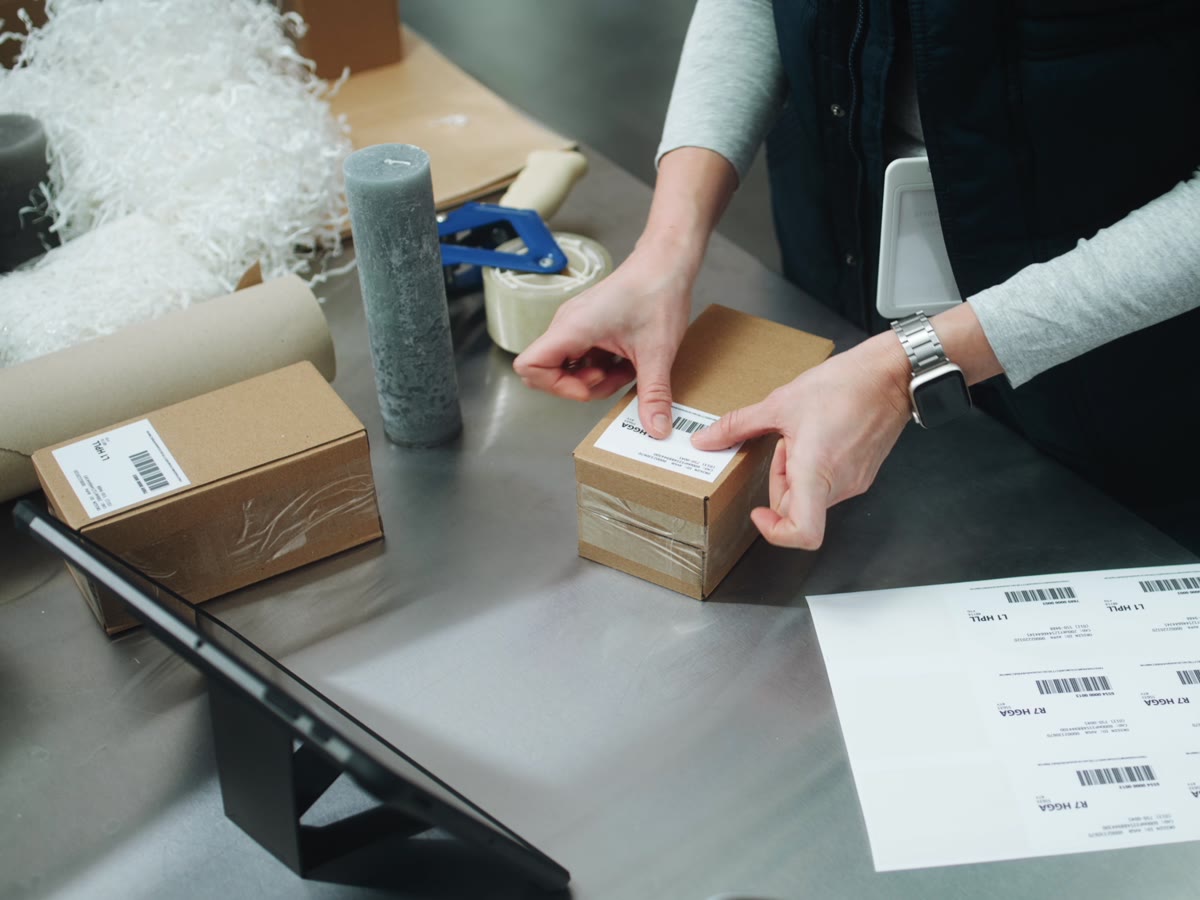 A Productiv operator applying a shipping label to a small D2C parcel at a pack station, with labels and dunnage ready